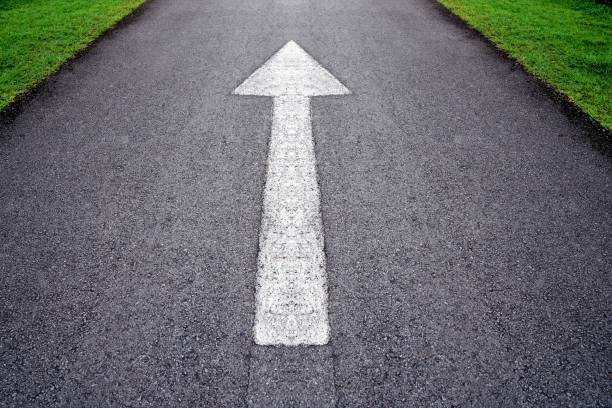 White forward arrow sign on grey asphalt road through the green grass field