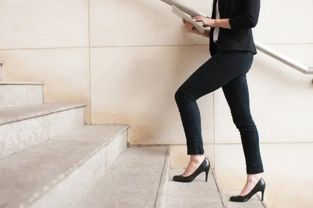 Portrait of young Caucasian businesswoman wearing high-heeled shoes walking upstairs and using digital tablet. Business concept