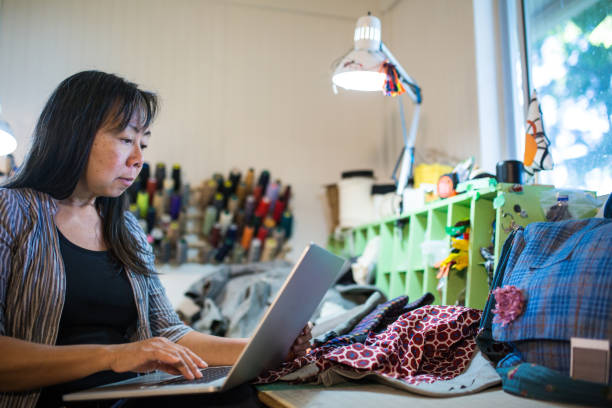 a mature textile entrepreneur working on a laptop computer in her studio - independence business women manual worker imagens e fotografias de stock