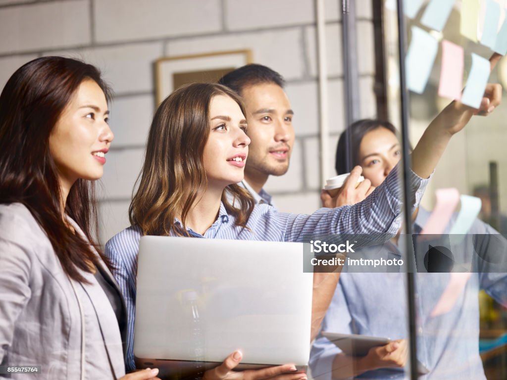 asian and caucasian business team working in office caucasian businesswoman pointing to a sticky note on glass wall during a brainstorming workshop in office. Challenge Stock Photo
