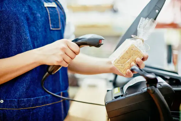 Midsection of saleswoman scanning barcode on food package in store. Young female deli owner is working at checkout counter. She is wearing denim overalls in shop.