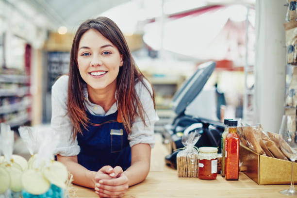 Portrait of confident deli owner leaning on checkout counter Portrait of confident deli owner leaning on checkout counter. Happy young saleswoman is in store. She is wearing denim overalls. convenience store photos stock pictures, royalty-free photos & images