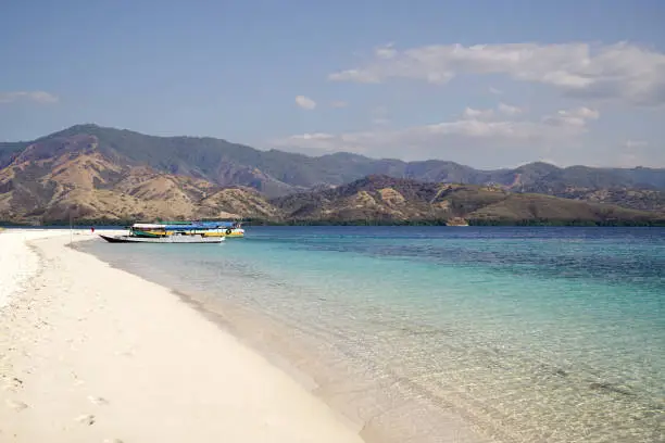 White sand beach with two boats on shore at midday overlooking broad island with green vegetation in the ocean, Flores Indonesia.