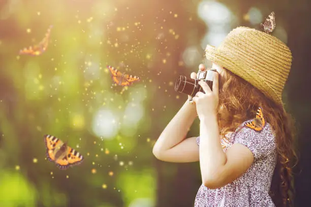 Photo of Little girl in straw hat, rustic style dress, photographing butterfly with retro photo camera in fairytale forest.