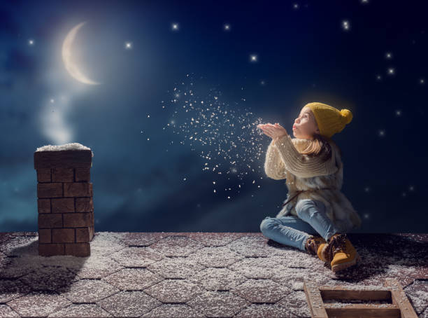 Happy child girl playing on a snowy winter walk. Little girl enjoys the game. Child girl sitting on the roof and looking at snowfall. Outdoor fun for winter vacation. Portrait kid on dark background.