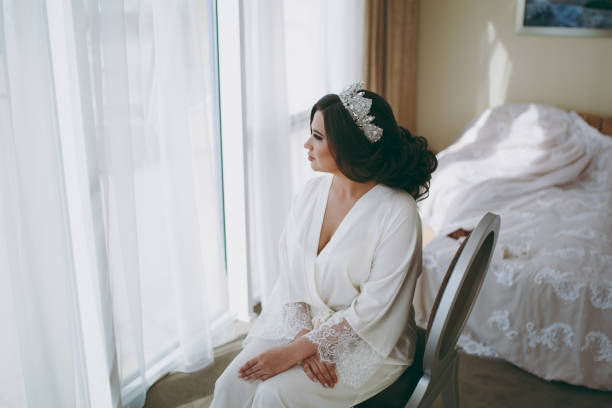 istock Bride brunette in white satin dressing gown sits by the window 855556872