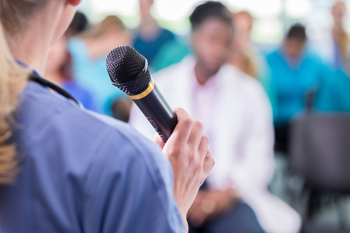 Unrecognizable female doctor speaks into a microphone as she addresses a group of doctors at a medical seminar or conference. Focus is on the microphone. The audience is blurred in the background.