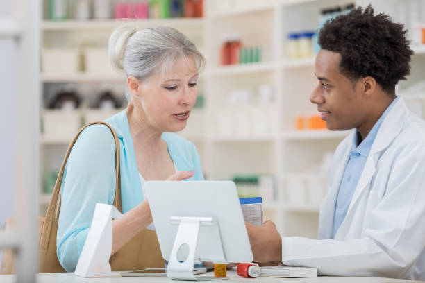 Senior woman asks pharmacy technician about cold medication A senior woman stands at the pharmacy counter with a male pharmacy technician and looks down at an over the counter boxed cold medication as she asks him about the side effects. older woman purse stock pictures, royalty-free photos & images