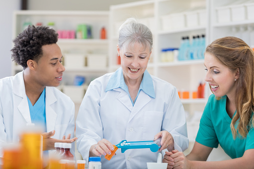 A senior female pharmacist stands at a table in her pharmacy with two attentive young coworkers and pours prescription tablets into a bottle as she explains each step in the compounding process.