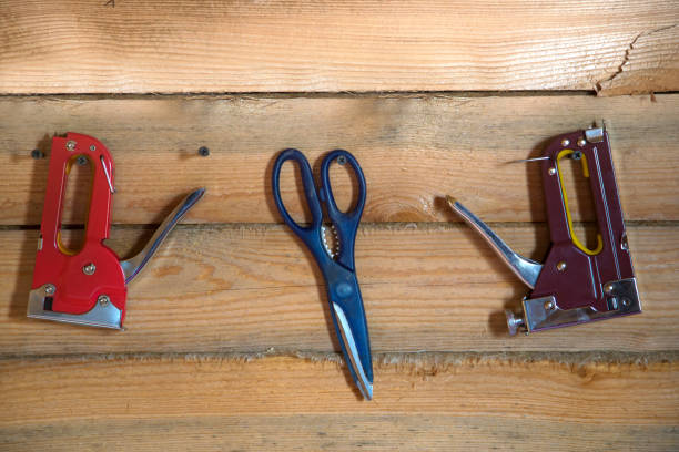 Tools for building on wooden background of pine forest. Staplers and scissors hanging on the wall on the screws. Between the pine rail laid with linen tape.