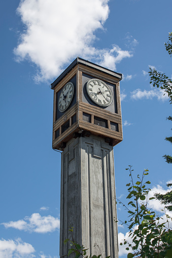 tall clock tower on the square of a small town in alaska during the summer time backed with a bright blue sky and white puffy clouds, made of wood and stone material showing the time on two sides of the top