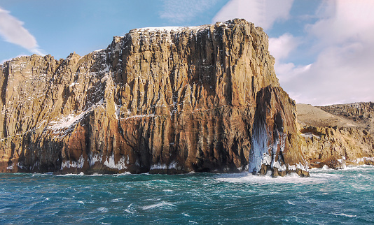 Close up detail of the steep cliff and sea stack with ice and frozen sea spray where the water hits the land.