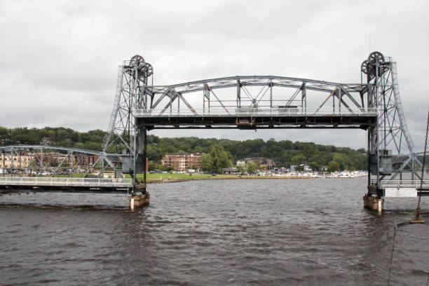 In Stillwater, Minnesota, this is an old lift bridge under construction, with a bright blue sky in the far background and a river lined with green trees, some construction machinery is sitting on the actual bridge and an orange sign shows the way to the channel