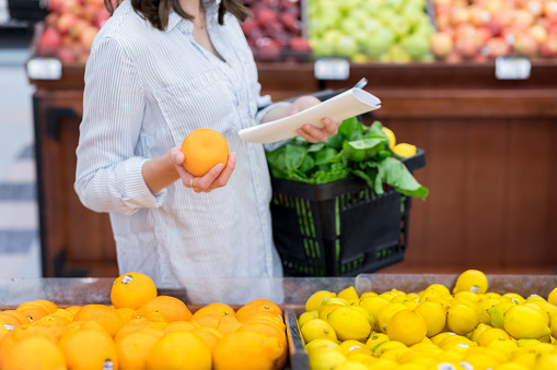 Unrecognizable woman shops for oranges in the produce section of a grocery store. She is squeezing the orange. She is holding a shopping basket filled with fresh produce.