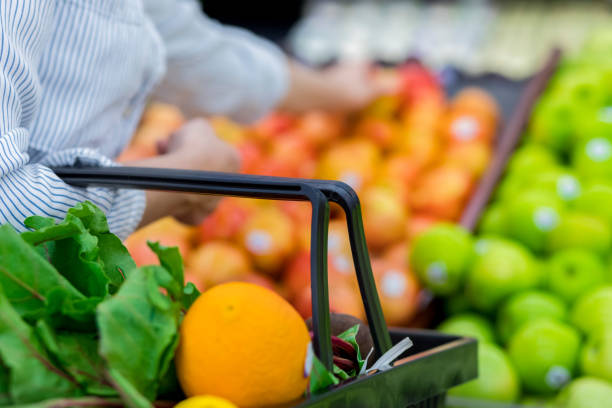 Unrecognizable female supermarket customer reaches for fresh oranges while shopping for produce. Focus is on a shopping basket filled with leafy greens and fresh oranges.