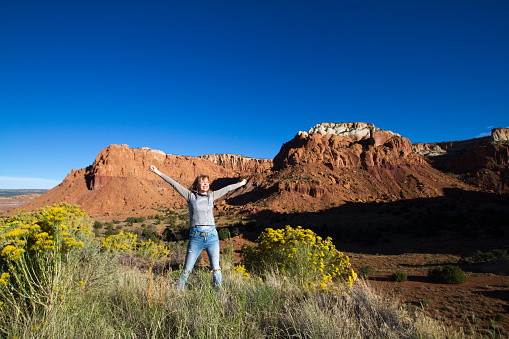 Wilderness victory: Senior Woman in early morning with arms raised in joy in front of red-orange rocks. Shot in Abiquiu, 50 miles north of Santa Fe, NM. Copy space in the vibrant blue sky.