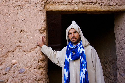 Zagora, Morocco: Portrait of a carpet seller in a burnoose stands outside his adobe doorway.