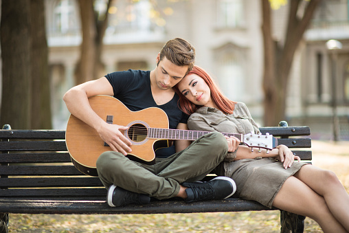 An attractive young man is seen sitting on a bench outside and playing an acoustic guitar while his girlfriend is sitting next to him and resting her head on his shoulder.