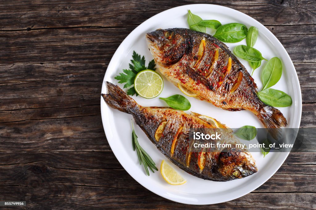 roasted sea bream fish with lemon slices delicious roasted dorado or sea bream fish with lemon and orange slices, spices, fresh parsley and spinach on white platter on old dark wooden table view from above Fish Stock Photo