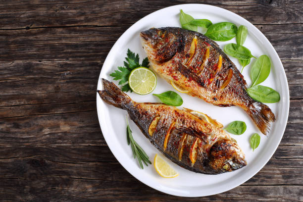 roasted sea bream fish with lemon slices delicious roasted dorado or sea bream fish with lemon and orange slices, spices, fresh parsley and spinach on white platter on old dark wooden table view from above roast stock pictures, royalty-free photos & images