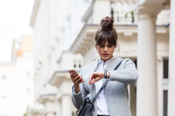 Elegant worried woman standing in front of city house in London, holding smart phone in hands and checking time. Autumn season.