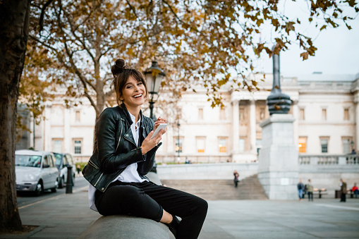 Beautiful, happy woman wearing leather jacket sitting at Trafalgar Square in London and using smart phone. Autumn season. National Gallery in the background.