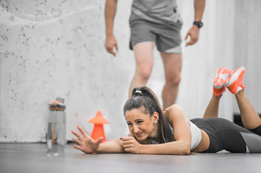 Young woman lying on the floor and doing new kind of exercise to stretch , she stretches to reach the bottle with water,and the fitness instructor is helping her with motivation