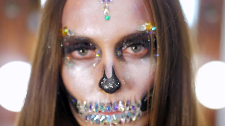 Scary young girl with creative halloween war-paint in the dressing room. Portrait of glamorous skull with rhinestones and sequins. Professional make-up for the celebration. Slow motion