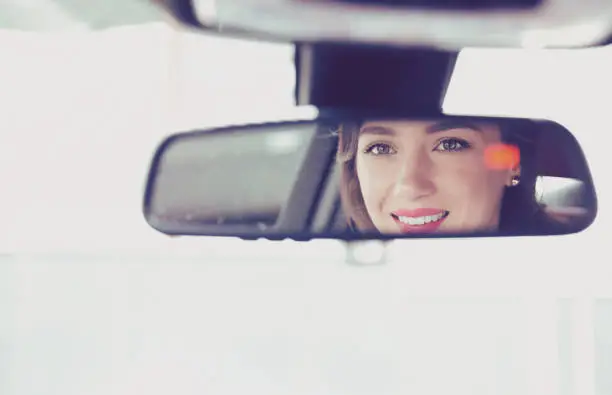 Young cheerful woman driving a car, rear view, reflexion of face in a mirror