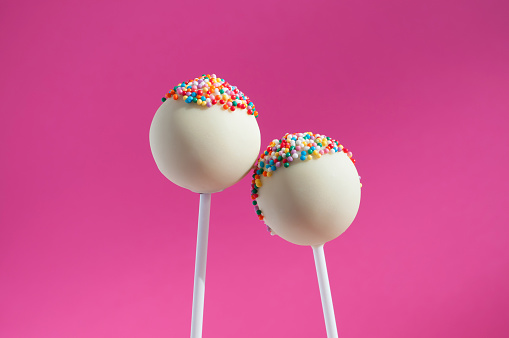 Two white chocolate cake pops decorated with colorfull confectionery sprinkles on a pink background. Concept sweet couple in love for your design. You can draw faces, emotions. Stick man.
