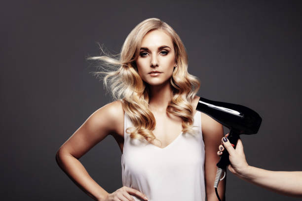 Gorgeous blond woman in a studio with hair dryer Portrait of young gorgeous blond woman standing on grey background with her hair being blown by dryer. blow drying stock pictures, royalty-free photos & images