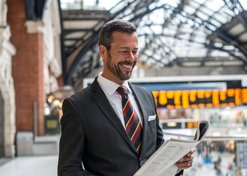 Man in full suit and with suitcase leaving the hotel, calling taxi and coming to the airport or train station, ready for his business trip. While waiting for check-in, man is drinking coffee and reading newspaper.