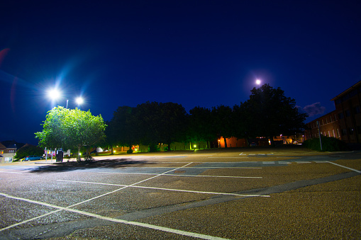 A public car park is illuminated by lights from a nearby building and the full moon creates silhouettes amongst the trees.