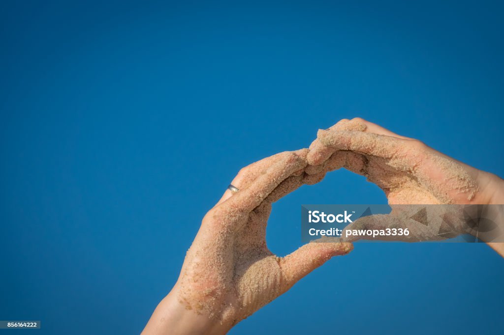 Hands covered in beach sand making a heart shape Close up of a sand covered woman hand making the heart shape with blue sky background Adult Stock Photo