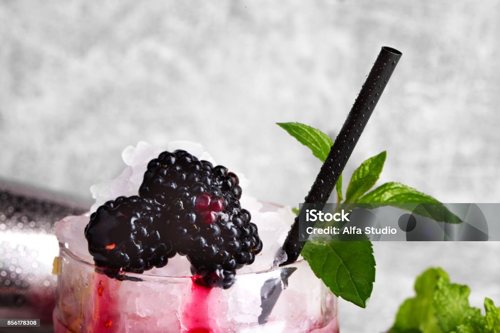 Close-up of a refreshing drink with mint twig and blackberries. A cocktail on a gray background. Healthy berries and mint. Macro picture of an icy berry beverage with mint leaves and blackberries on a gray background. Ripe, fresh blackberries and a black straw for a cold smoothie. Celebration concept. Copy space. Antioxidant Stock Photo