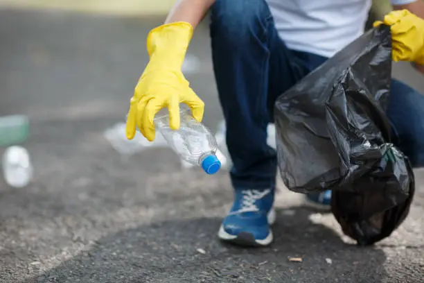 Photo of Male hands in yellow rubber gloves putting household waste into small and black bin bag outside.