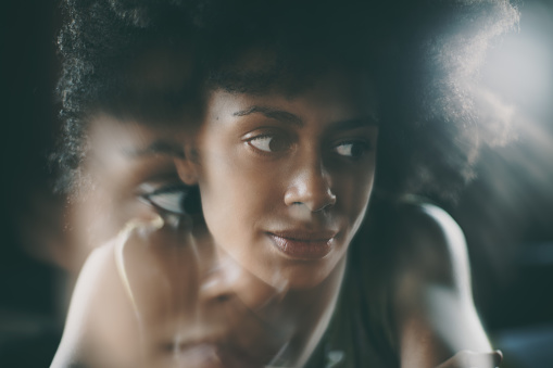 Double exposure close-up portrait of beautiful young biracial girl with curly arfo hair thoughtfully looking aside while sitting inside of dark place with light in background; shallow depth of field