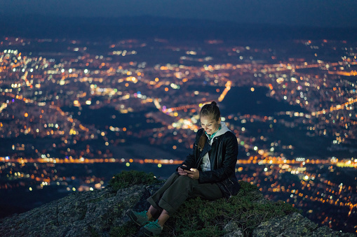 Young smiling woman texting on her mobile phone and sitting on the mountain peak after sunset. The lights of a big city lying in the valley below.