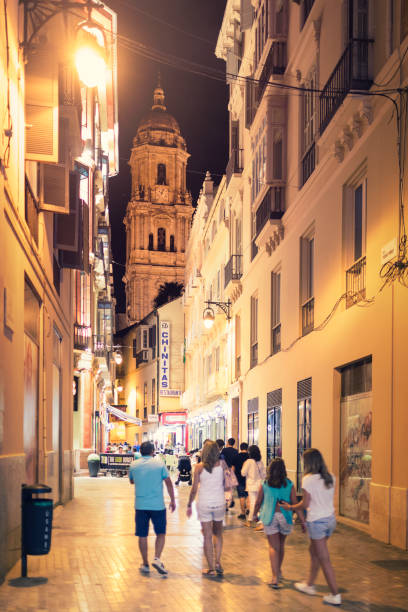 A group of people out walking through the old town streets of Málaga in the evening. In the background is the tower of Málaga Cathedral.