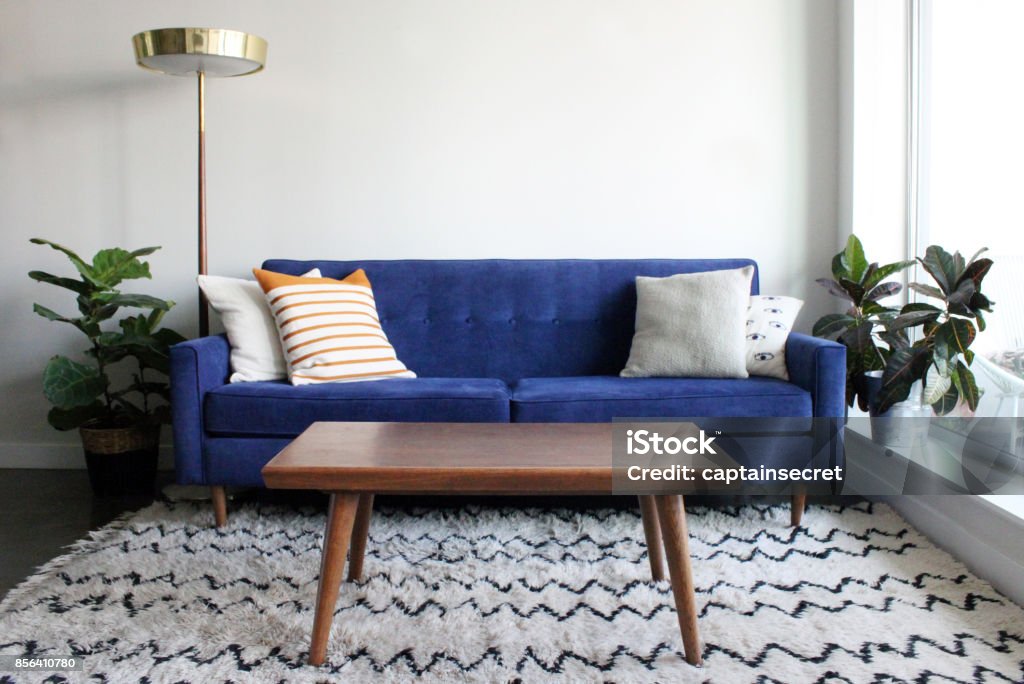 Blue Suede Mid Century Modern Couch in Minimalist Apartment Setting Low angle shot of a royal blue suede mid century modern 4 seater sofa and teak coffee table. Set in a modern apartment living room complete with mid century furnishings, moroccan rug and potted plants. Coffee Table Stock Photo