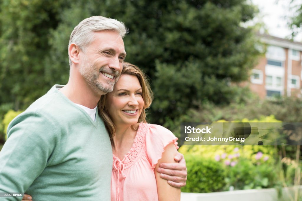 Mature man with arm around woman and looking away smiling Cheerful couple in garden, woman leaning against man, trees and shrubs in background 50-54 Years Stock Photo