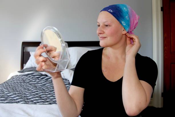 Young cancer patient tries on a new headscarf in the mirror and smiles stock photo