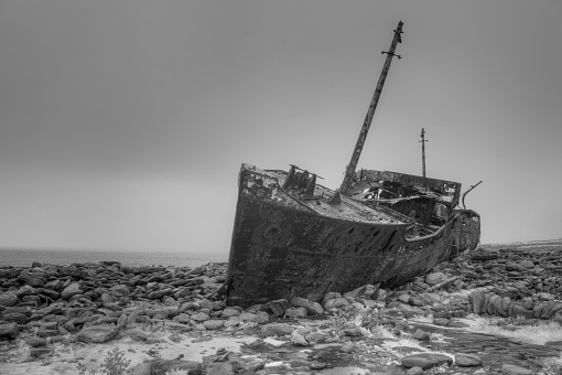 A black and white image of the twisted, rusting hulk of a decades-old shipwreck on the shores of Inisheer (Inis Oirr) in the Aran Islands, Ireland. Shot with the ocean in the background, behind the ship, using an infrared camera.