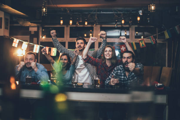 Celebrating in the pub Group of young people, sitting in a pub all together, watching a sports game. spectator stock pictures, royalty-free photos & images