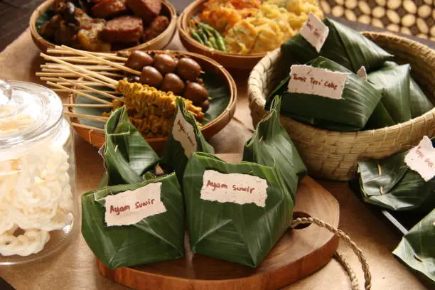 This is the luxury version of Angkringan Nasi Kucing, a popular street food fare of small portion of rice dish in banana leaf package (a portion so small resembling a cat's rice portion, hence the name nasi (rice) kucing (cat)). The rice dish is topped with variety of toppings such as shredded chicken, sautéed tempeh, and sautéed anchovy. Each portion dish is wrapped in banana leaf and grouped together based on the topping. The package is given handwritten label of the topping inside. The rice dish are accompanied with extra side dishes of satays (chicken intestine, quail eggs, cockles), sweet-soy braised food (tofu, tempeh, offal) and fritters (vegetables and tempeh). Some crackers are also provided to complete the meal. The food are arranged on a wooden table lined with recycled papers.