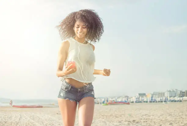 african curly hair girl running on beach listening to music with smart phone