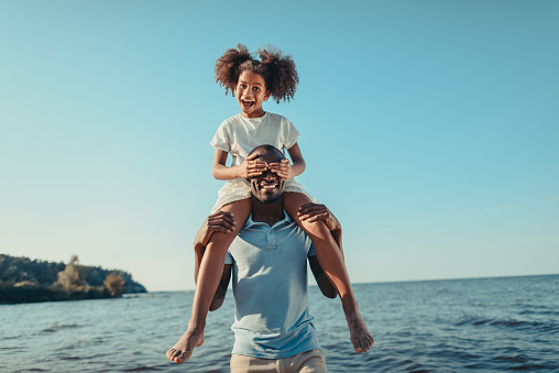 smiling african american father carrying adorable little daughter on beach