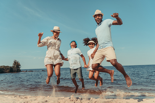 happy african american family holding hands and jumping on beach