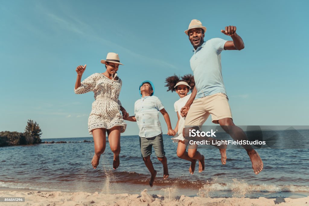 family on beach happy african american family holding hands and jumping on beach Family Stock Photo