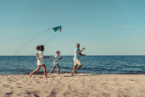happy african american father and kids playing with kite on beach